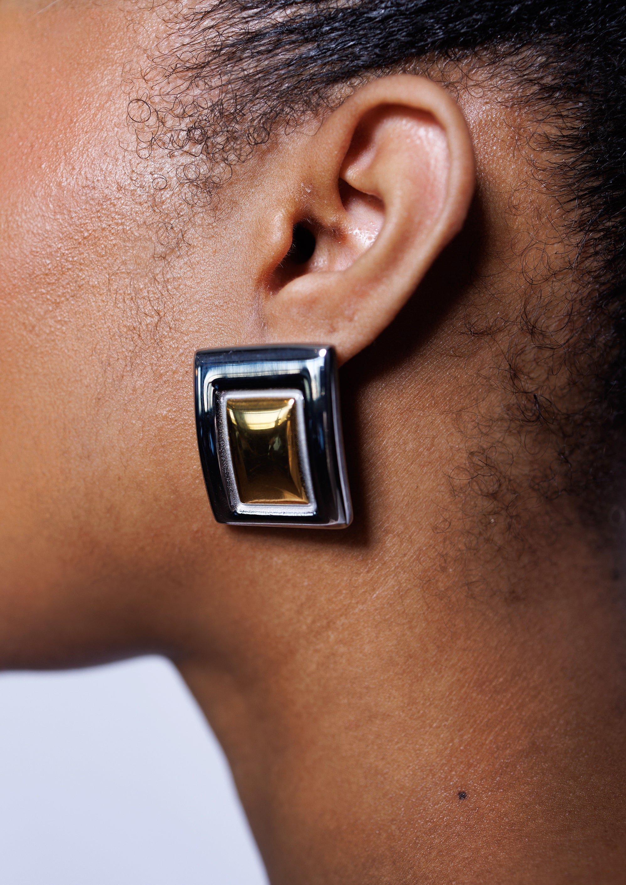 Close-up of an ear wearing a gold and black rectangular earring against a neutral background