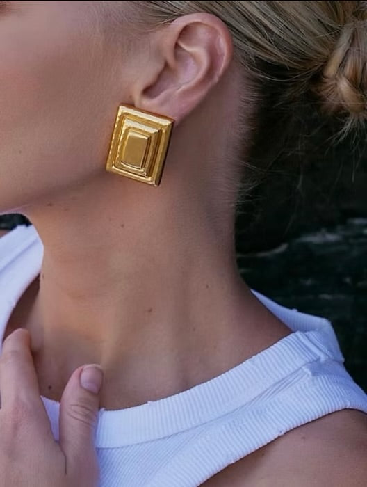 Gold square earrings worn by a person with a blurred background