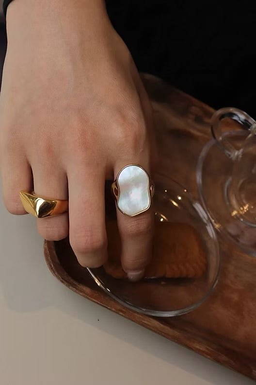 Hand wearing a gold ring with a large pearl-like stone, holding a glass container.