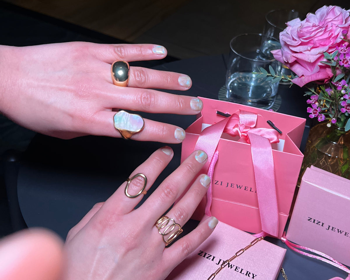 Hands wearing rings with a pink jewelry box and flowers on a table