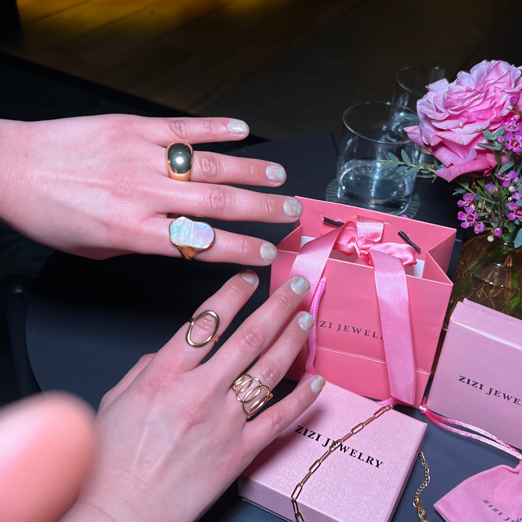 Hands wearing rings with a pink jewelry box and flowers on a table