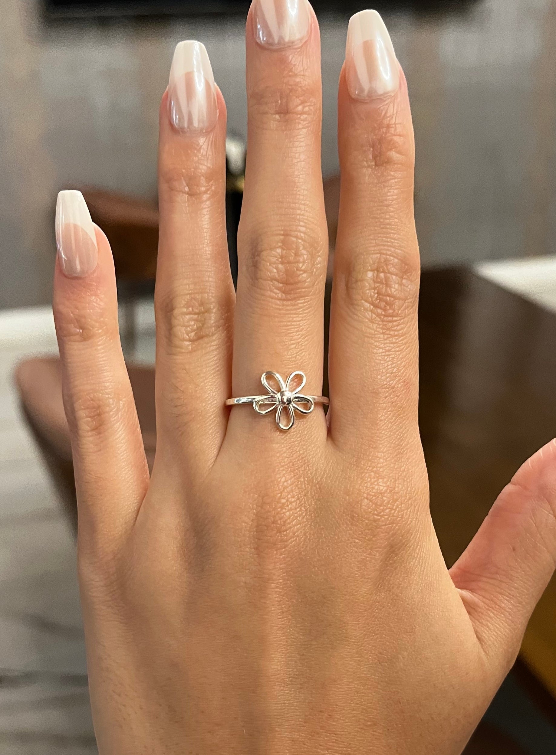 Hand wearing a silver ring with a flower design, set against a blurred indoor background.