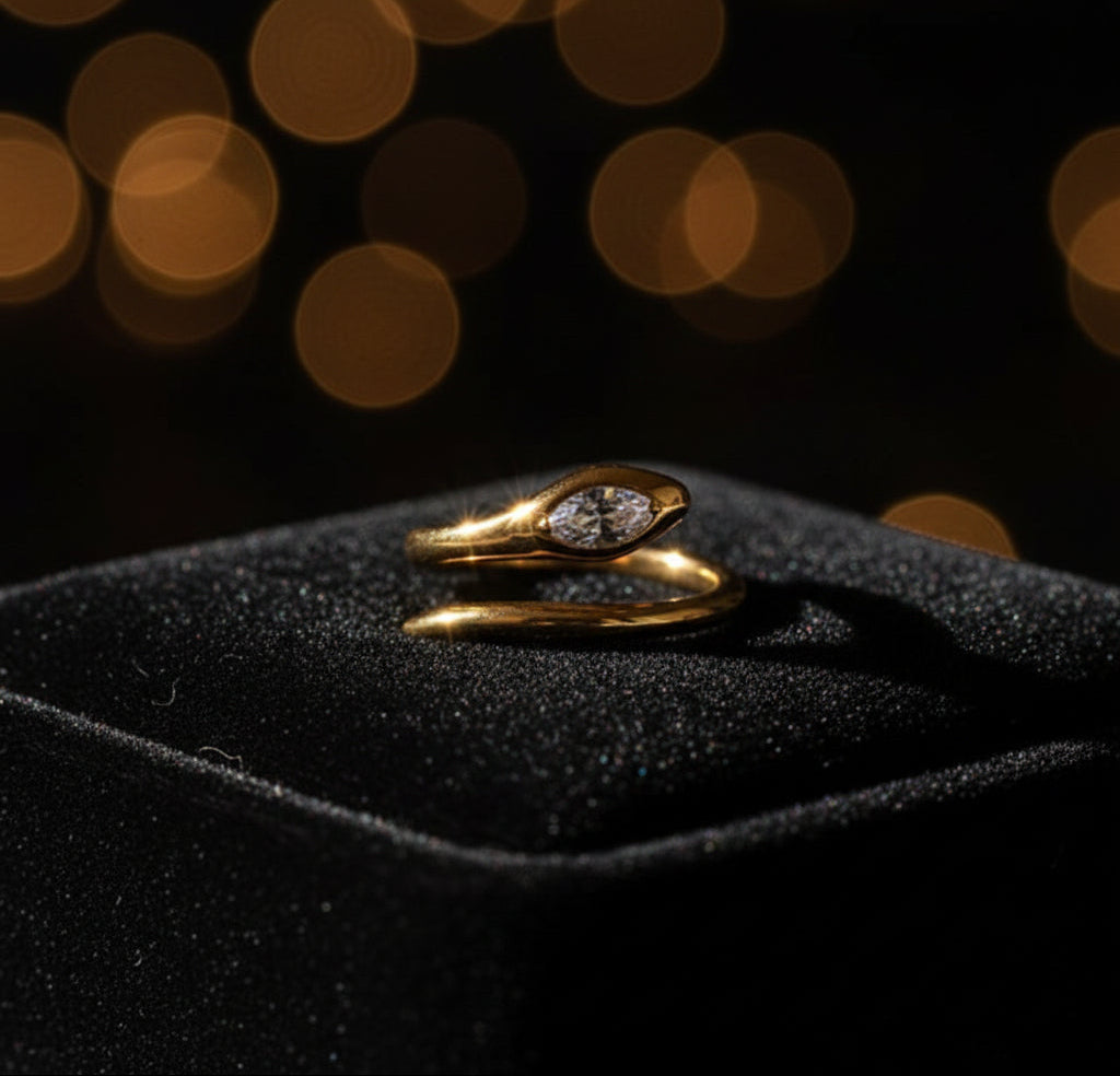 A gold snake ring with a clear teardrop crystal, resting on a textured beige stone surface.
