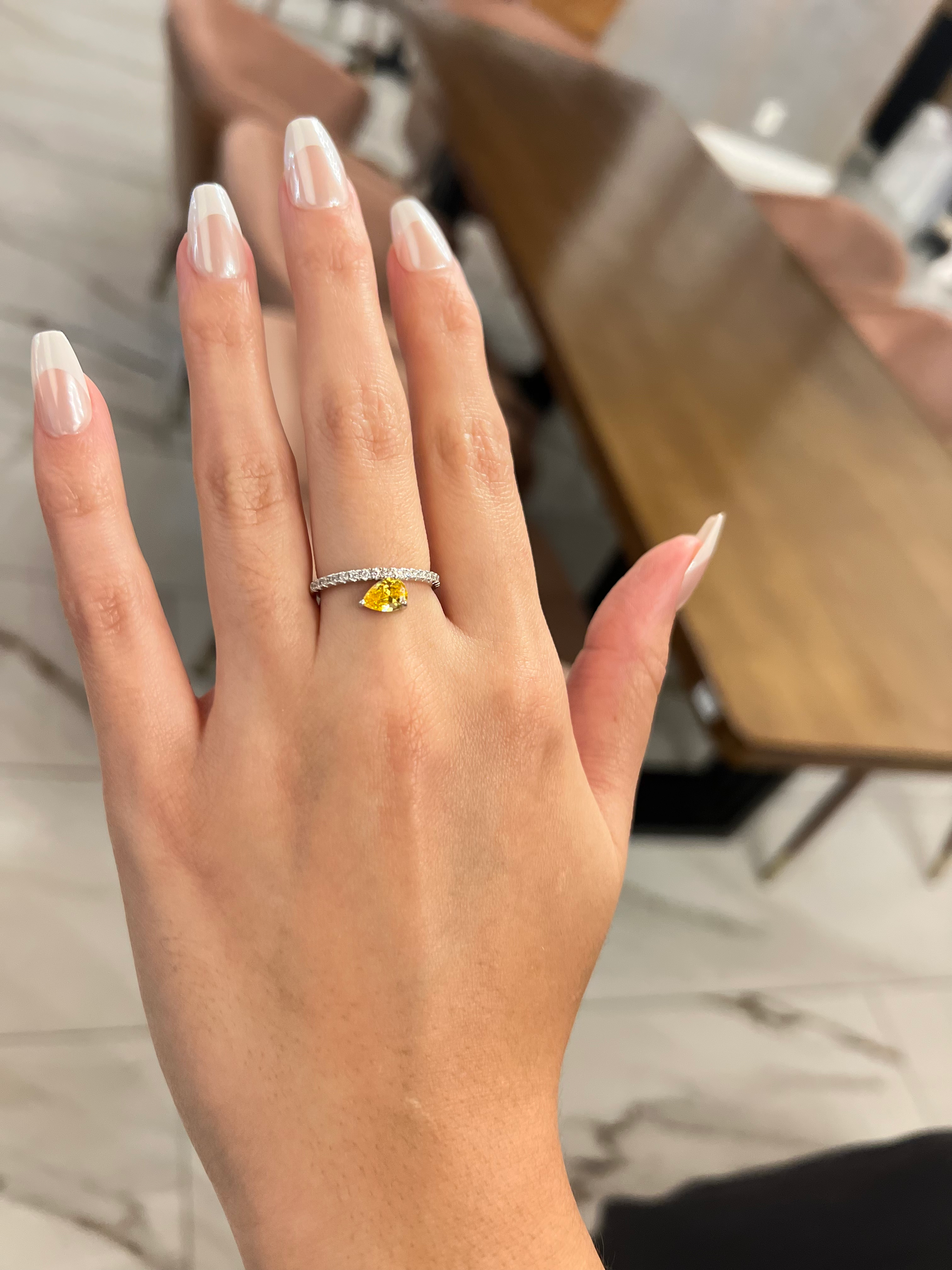 Hand wearing a ring with a yellow gemstone against a blurred indoor background