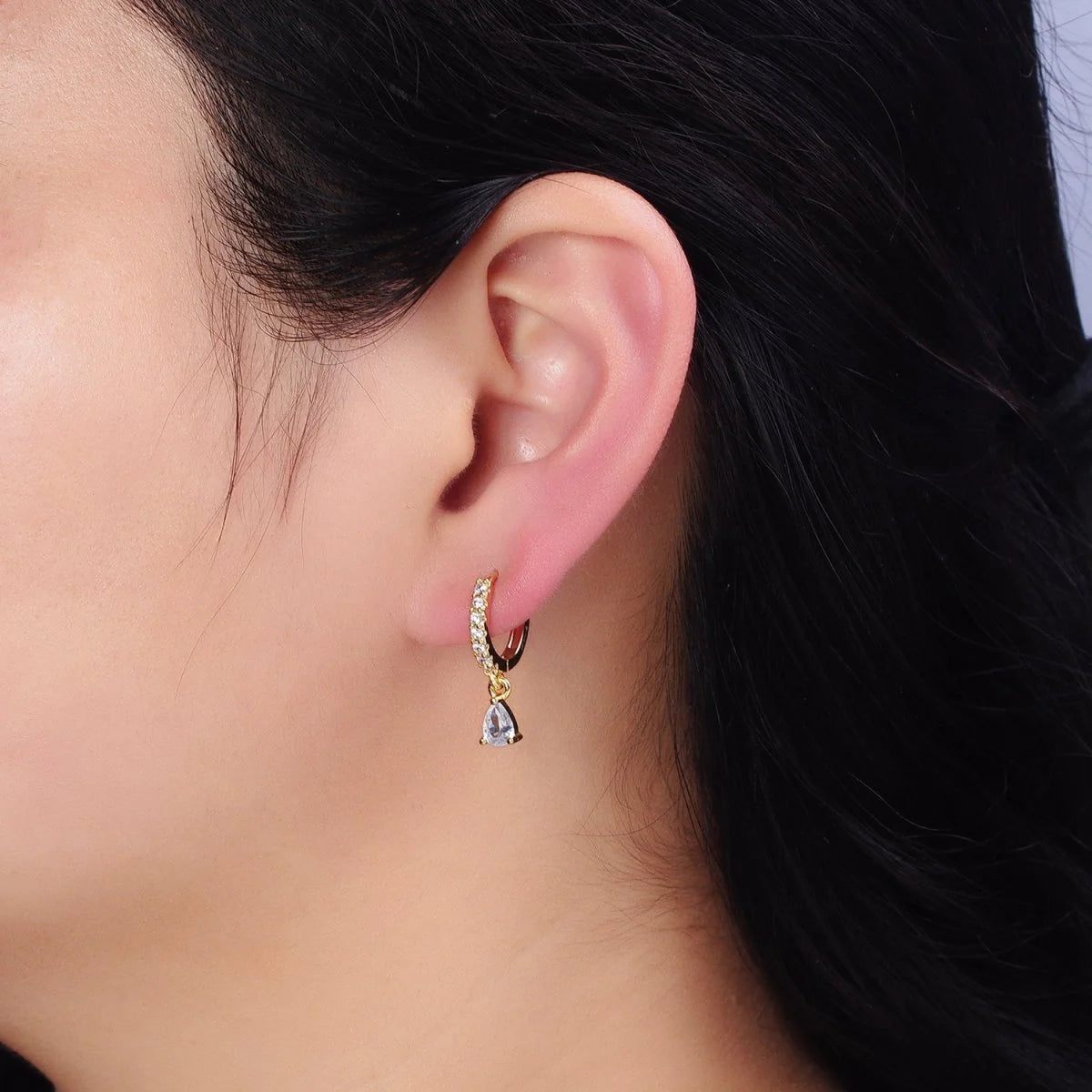 A close-up of a person's ear wearing a small gold huggie hoop earring with a dangling clear teardrop crystal charm.