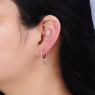A close-up of a person's ear wearing a small gold huggie hoop earring with a dangling clear teardrop crystal charm.