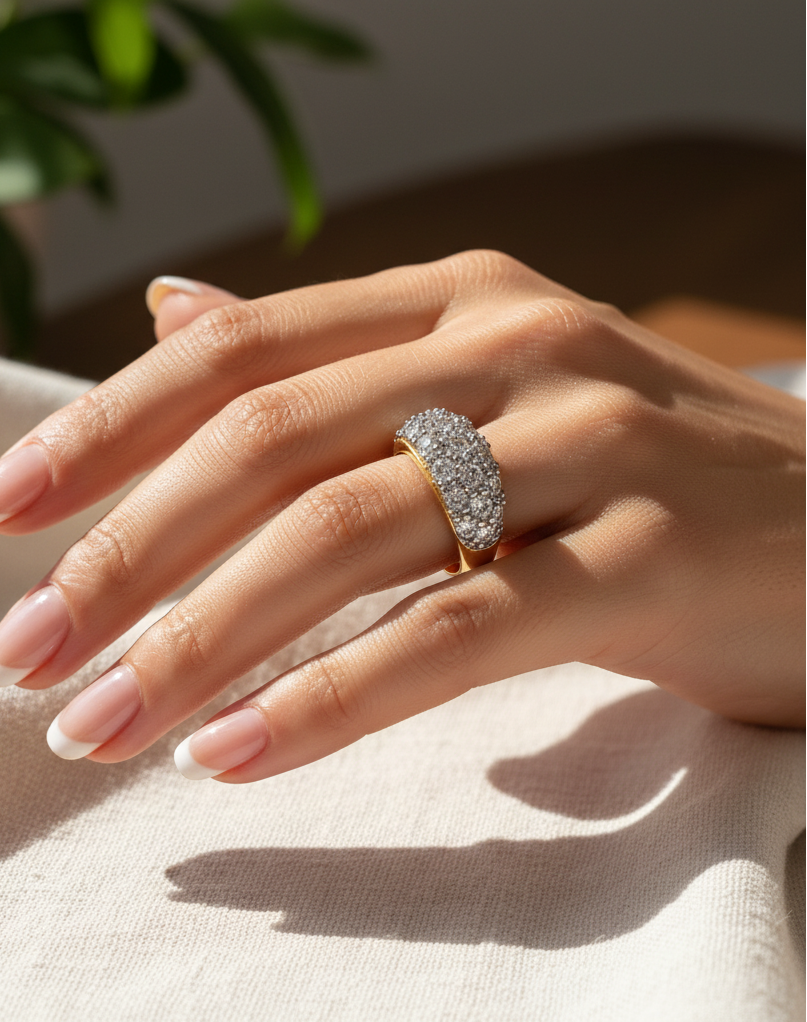 A close-up of a hand wearing a wide gold ring set with clear pavé stones, resting on a light beige fabric surface with a soft-focus background.