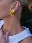 Gold square earrings worn by a person with a blurred background