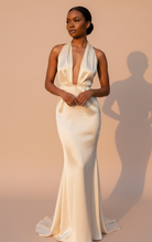 A woman stands in a studio wearing a satin halter-neck mermaid gown in a warm ivory or cream tone. The dress has a deep V-neckline, fitted silhouette, and a slight train.