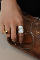 Hand wearing a gold ring with a large pearl-like stone, holding a glass container.