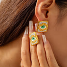 Gold and green eye-shaped earrings worn by a person with brown hair.