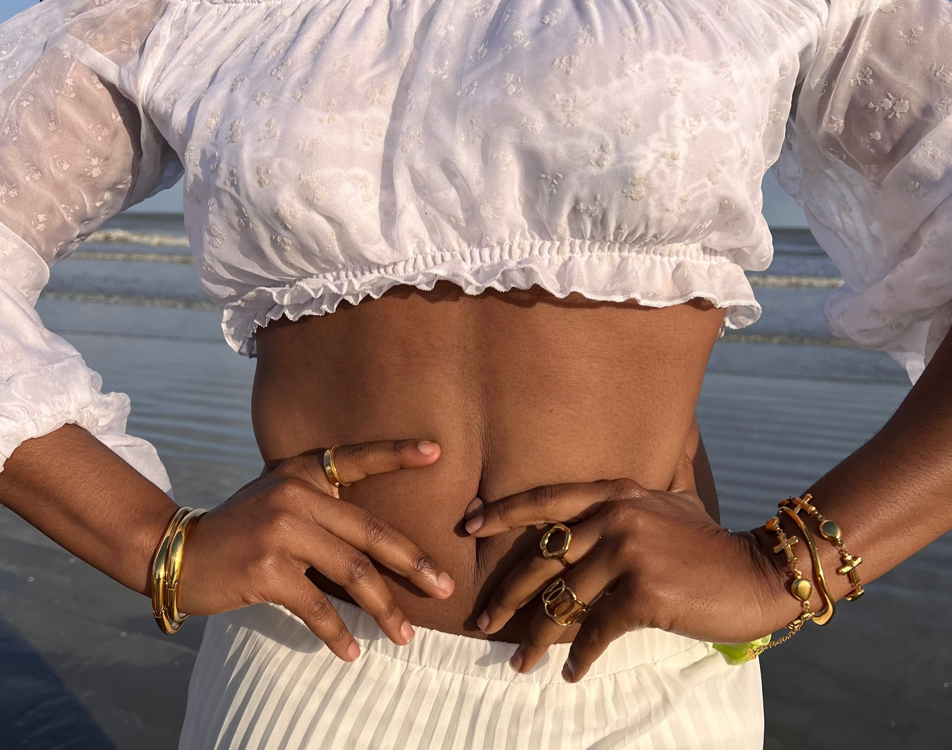 Person wearing a white off-shoulder top and skirt on a beach with gold bracelets, gold rings, and colorful charm necklaces