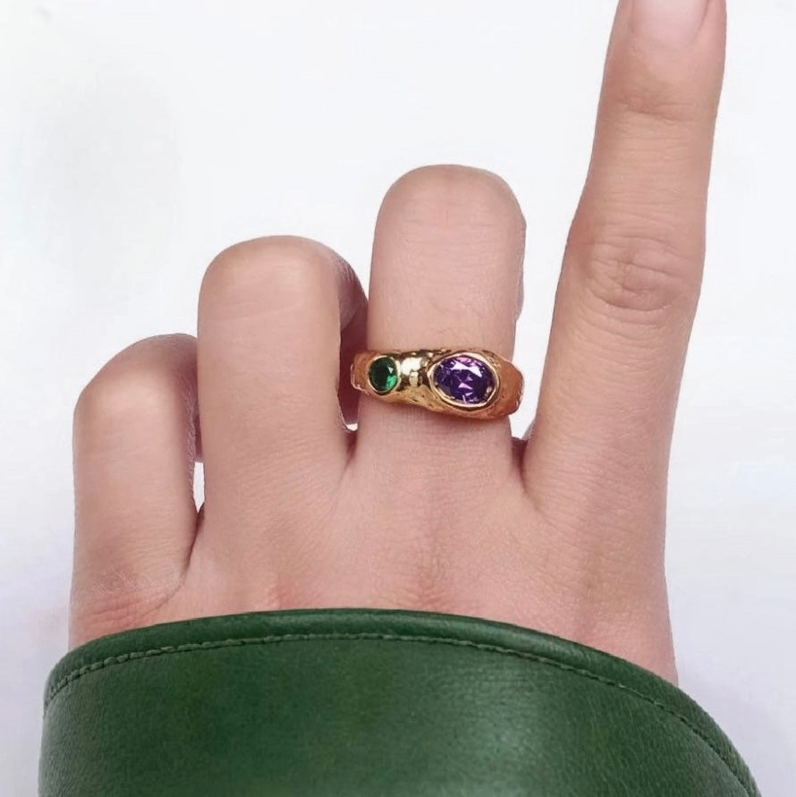 Gold ring with green and purple gemstones on a finger against a white background