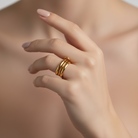 A close-up of a hand wearing three slim, parallel gold rings on one finger, set against a softly blurred warm beige background.