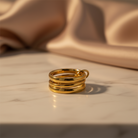 A gold ring with three stacked, parallel bands and a small loop at the top, resting on a light marble surface with draped satin fabric in the background.