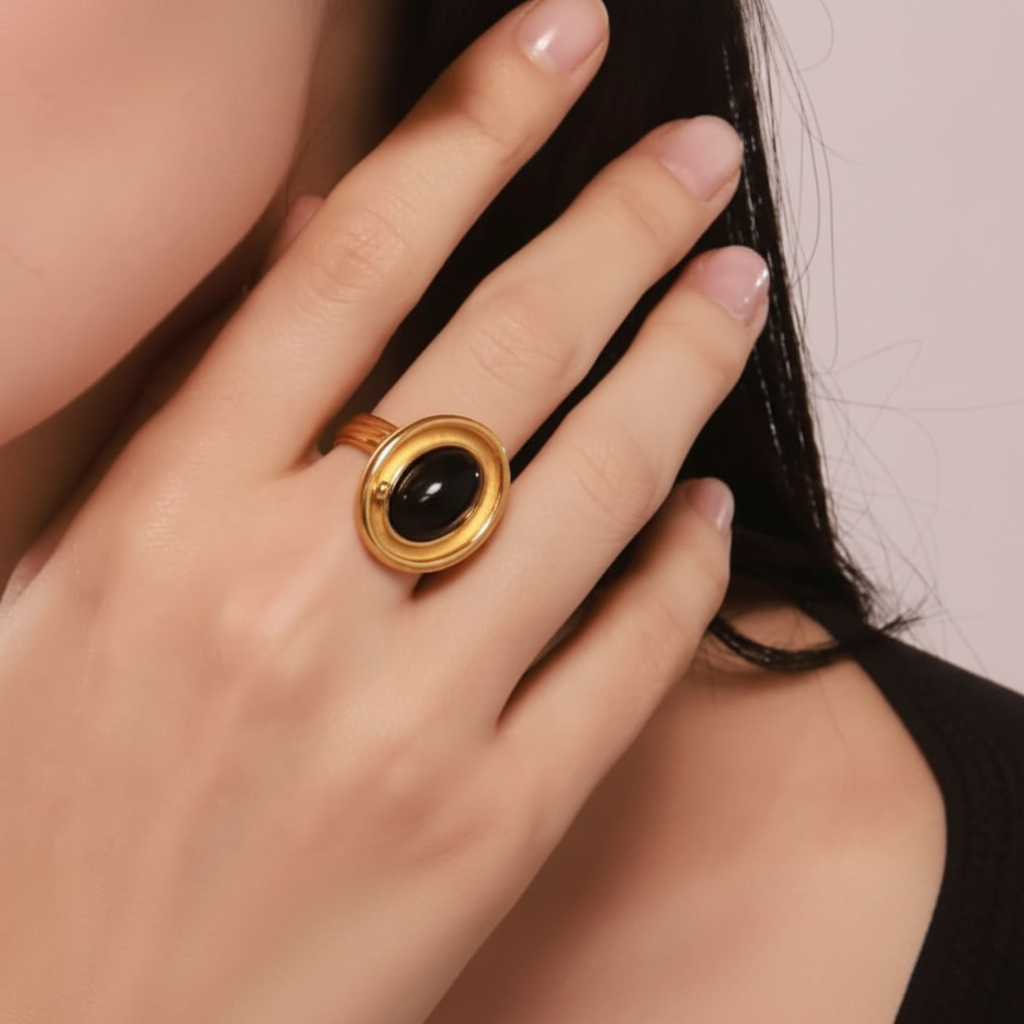 A close-up of a model wearing a gold ring and matching stud earring, both featuring oval black cabochon stones in gold bezel settings.