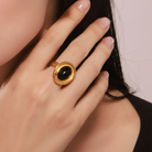 A close-up of a model wearing a gold ring and matching stud earring, both featuring oval black cabochon stones in gold bezel settings.