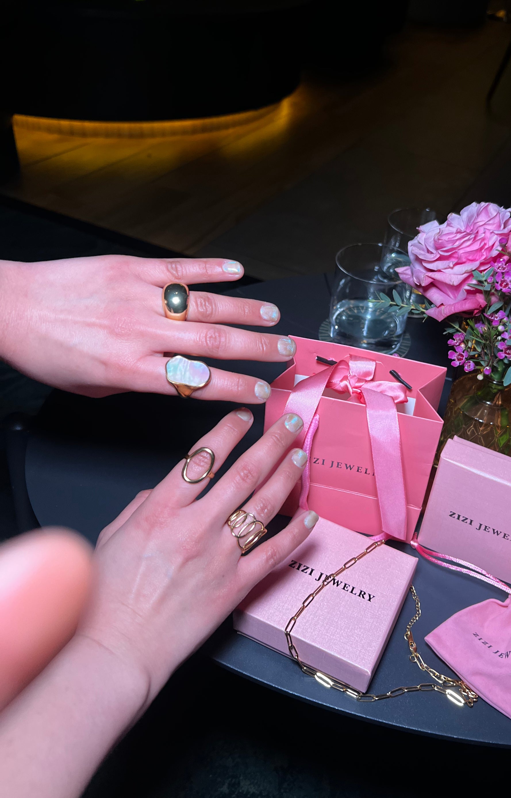 Hands wearing rings with a pink jewelry box and flowers on a table
