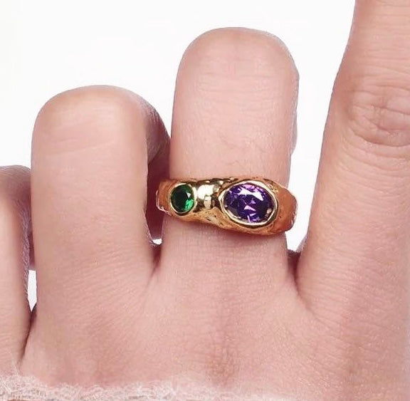 Gold ring with green and purple gemstones on a finger against a white background