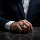 Hand wearing a ring on a dark surface with a suit jacket in the background