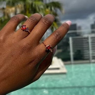 Hand wearing two red gemstone rings with a blurred pool and building background