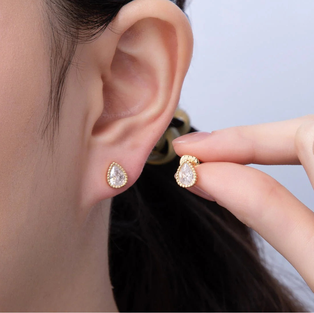 Gold teardrop earrings being worn and held up to an ear against a neutral background