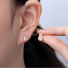 Gold teardrop earrings being worn and held up to an ear against a neutral background
