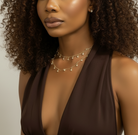 A close-up of a model wearing a double-layered gold necklace with small pearl-like beads and dangling charms, paired with a dark brown halter top.