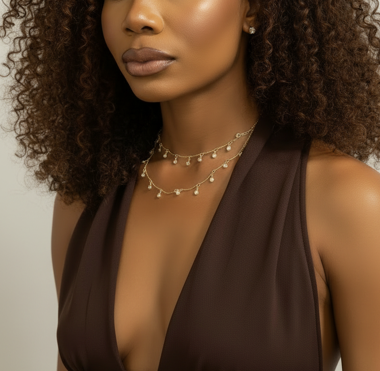 A close-up of a model wearing a double-layered gold necklace with small pearl-like beads and dangling charms, paired with a dark brown halter top.