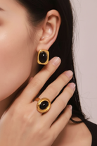 A close-up of a model wearing a gold ring and matching stud earring, both featuring oval black cabochon stones in gold bezel settings.