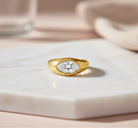 A gold signet-style ring with a marquise-shaped clear gemstone, resting on a white marble slab with a soft beige background.