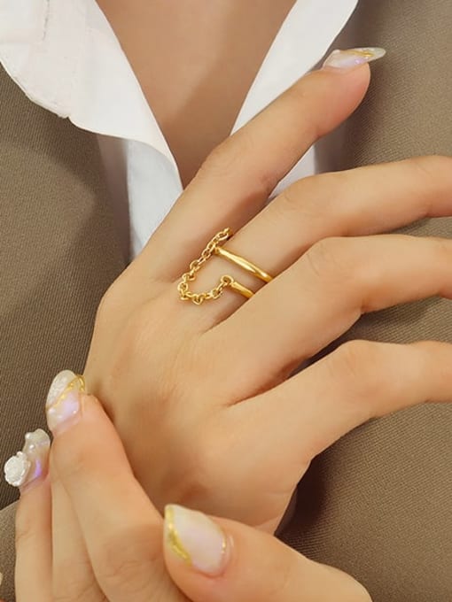 A close-up of a person's hand wearing a delicate gold ring with a short chain segment, styled with a white collared shirt and brown blazer.