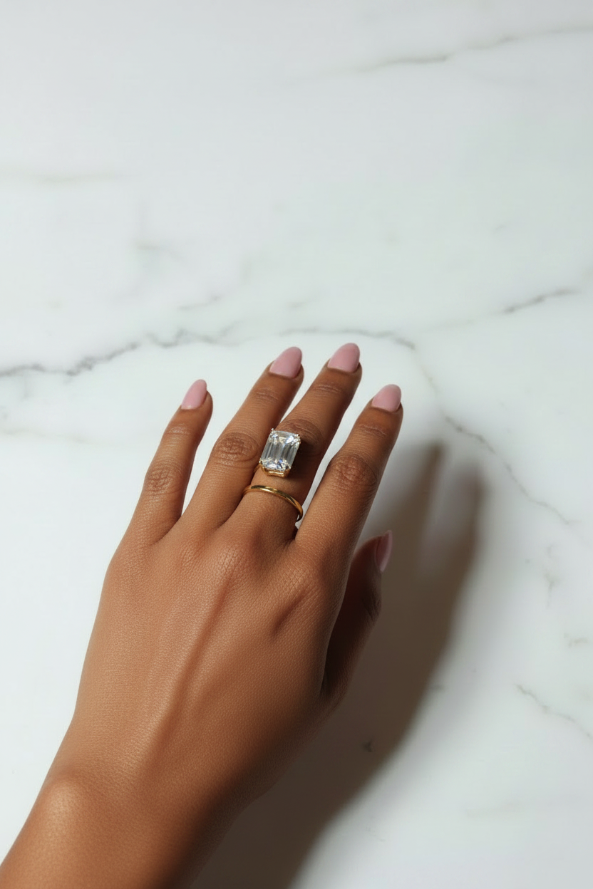 A hand with pink manicured nails wearing a silver-toned ring with a rectangular clear gemstone, resting on a white marble surface.