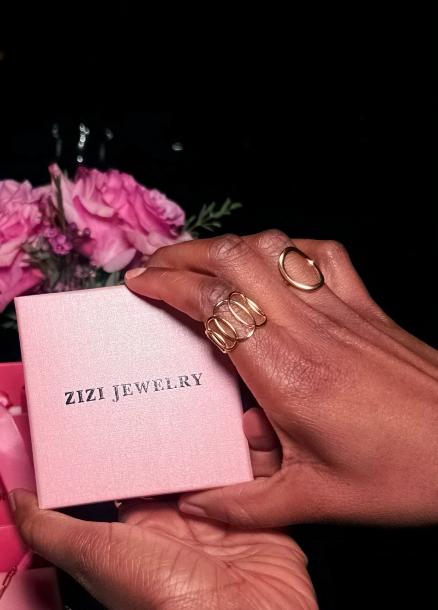 Hand holding a pink ZIZI JEWELRY box with a gold ring on a dark background