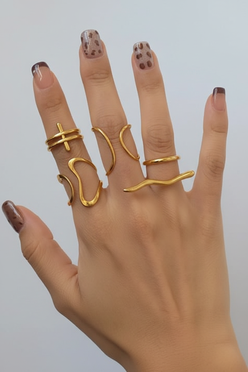 A hand with manicured nails wearing five thin gold rings in different abstract and wavy designs, set against a plain light gray background.