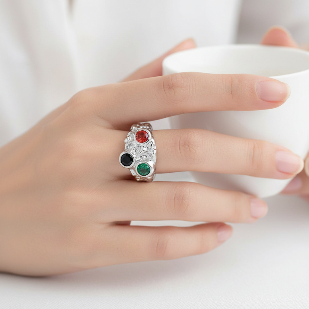 Hand wearing a ring with colored stones, holding a white cup against a neutral background