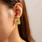 Close-up of an ear wearing a gold square earring with a green eye design.