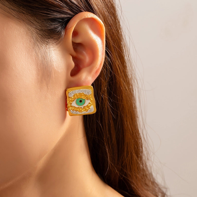 Close-up of an ear wearing a gold square earring with a green eye design.