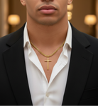 Man wearing a black suit and white shirt with a gold necklace featuring a cross, set against a blurred indoor background.